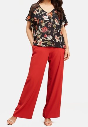 Femme portant un chemisier noir à manches courtes avec des motifs floraux et des détails en dentelle, ainsi qu'un pantalon rouge taille haute à jambes larges, se tenant les mains dans les poches.