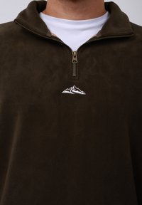 Pull-over beige en polaire avec un col montant, doté d'une fermeture éclair légère et d'un logo de montagne blanc brodé sur la poitrine.