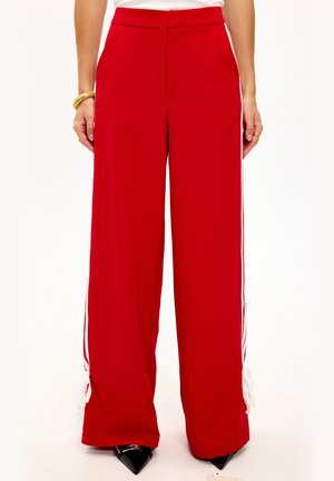 Pantalon large rouge avec des rayures blanches sur les côtés, présentant une texture lisse et des détails de nœud élaborés à l'ourlet. Talons aiguilles noirs visibles.