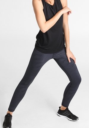 Débardeur de sport noir associé à des leggings sombres à motifs, avec une coupe ajustée et une surface texturée, porté avec des chaussures de sport noires.