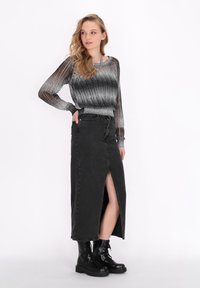 Fustă maxi din denim negru cu fante în față, purtată împreună cu un top cu mâneci lungi în nuanțe de gri și negru. Modelul poartă cizme negre cu șireturi.