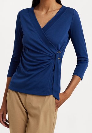 Vrouw draagt een blauw wikkelstijl langarmtop met een zijgesp, gecombineerd met beige broek, poserend met één hand in de zak.