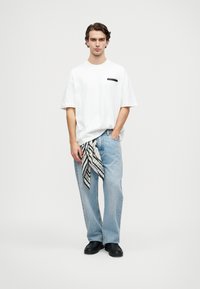 Jauns vīrietis stāv, ģērbies baltā pārāk lielā T-kreklā ar kabatas tekstu, gaismas zilos brīvos džinsos, melnās kurpēs un melnbaltajā šallē, kas karājas no kabatas.