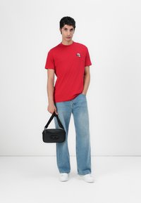 Raudona medvilninė marškinė su maža grafika ant krūtinės, derinama su šviesiai mėlynomis plačiakelnėmis džinsomis ir juoda petine rankine. Baltos sportbačiai.