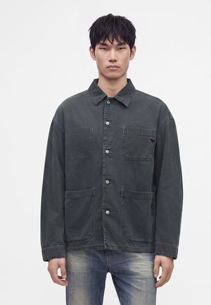 Junger Mann mit kurzen schwarzen Haaren trägt eine dunkelgraue, knöpfbare Jacke mit drei vorderen Taschen und ausgewaschene blaue Jeans vor einem einfarbigen Hintergrund.