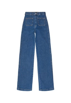 Femme portant un jean bleu taille haute à jambe droite, un haut blanc et des chaussures à plateforme noires, debout, dos à la caméra sur fond blanc.