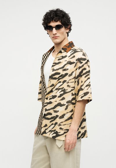 Man met krullend haar, zwarte zonnebril, een overhemd met half ruitjes-/half dierenprint, wit ondershirt en beige broek, zijwaarts poseerend.