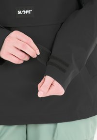 Musta takki, jossa on SLOPE-logo, tarranauhalla säädettävät hihansuut ja kolme vaakasuoraa raitaa. Liukas materiaali ja istuva muotoilu.