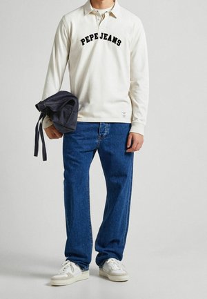 Homme portant une chemise blanche à manches longues avec le texte "PEPE JEANS", un jean bleu, des baskets blanches, tenant une veste en denim repliée dans la main gauche.