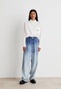 Λευκό πουκάμισο με μακρύ μανίκι, τσέπη στο στήθος και λογότυπο, σε συνδυασ�μό με φαρδιά, ξεθωριασμένα ανοιχτό μπλε τζιν και χοντρές μαύρες μπότες.