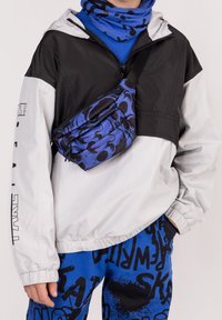 Czarna i biała kurtka przeciwwiatrowa z pół-zamkiem, niebieskie dresy z grafiką oraz niebieska i czarna torba na ramię w wzory. Gładkie, nowoczesne tekstury.