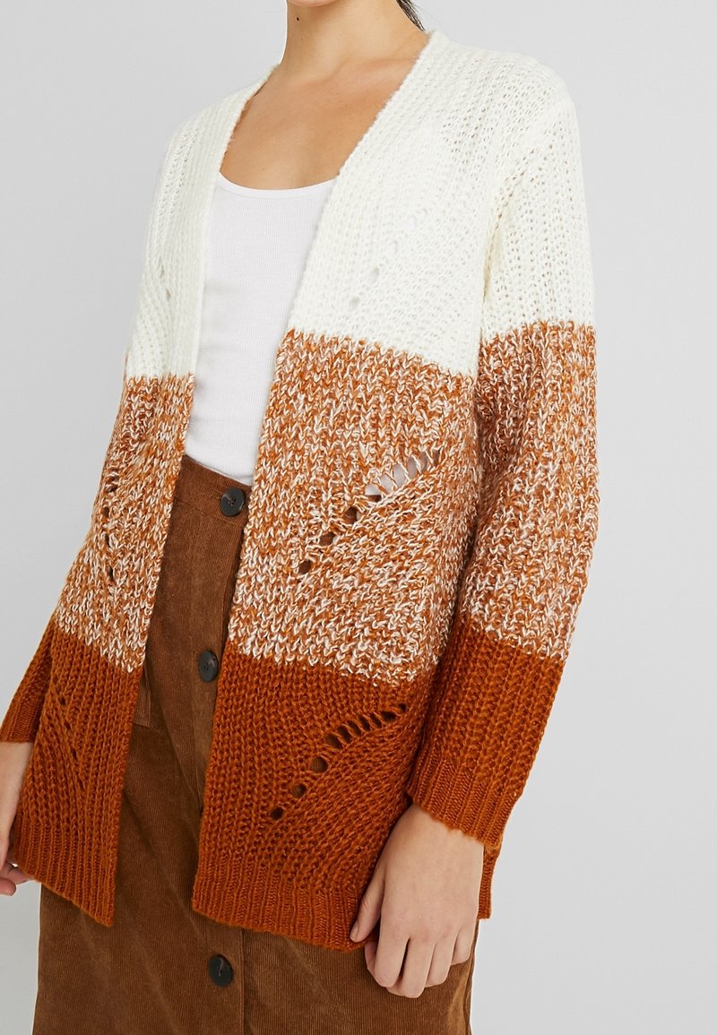 Femme portant un cardigan en tricot tricolore blanc, beige et rouille, par-dessus un débardeur blanc et une jupe boutonnée marron.