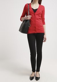 Röd kofta med V-hals och knappar, kombinerad med en svart topp, smala svarta byxor och svarta lägenheter. Bär en svart handväska med kedja.