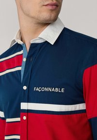 Camisa abotonada en azul marino, rojo y blanco. Presenta rayas horizontales, un cuello blanco y un logo bordado en el pecho. Material de mezcla de algodón.