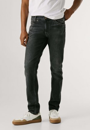 Mand iført falmede sorte slim-fit jeans, hvide sneakers med brune såler og en hvid t-shirt, stående med den ene hånd på hoften.