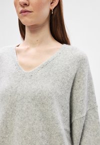 Suéter tricotado cinza claro com decote em V, ombros caídos e uma textura suave. Apresenta pequenos detalhes tricotados ao longo do decote.