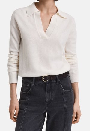 Frau trägt einen langärmeligen, elfenbeinfarbenen Pullover mit V-Ausschnitt, der in hochtaillierte schwarze Jeans mit schwarzem Gürtel gesteckt ist.