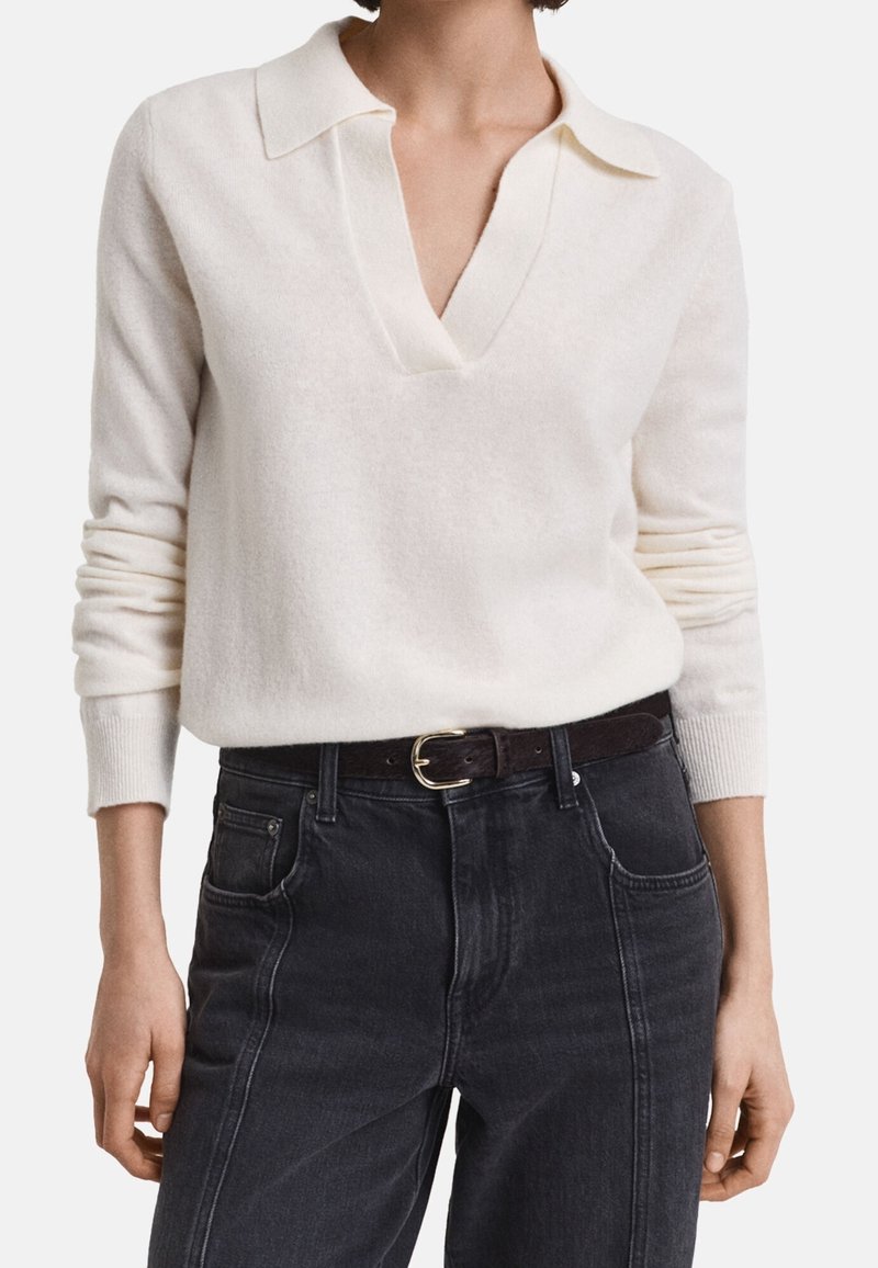 Femme portant un pull à manches longues écru avec un col en V, rentré dans un jean noir taille haute avec une ceinture noire.