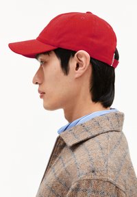 Casquette de baseball rouge en tissu, avec une visière incurvée et une sangle ajustable. Portée par une personne aux cheveux noirs, associée à une veste à motifs.