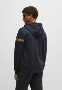 Marinblå hoodie med en gyllene rand på vänster axel, har en avslappnad passform och mjuk textur. Matchas med marinblå byxor.