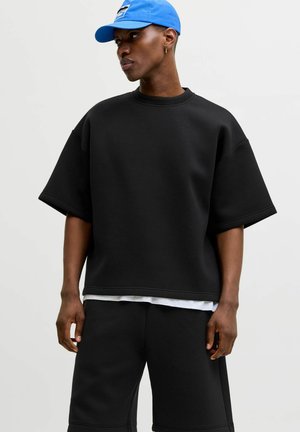 Osoba ubrana w luźną czarną koszulkę z krótkim rękawem i czarne szorty, widoczna biała podkoszulka oraz niebieska czapka z daszkiem, patrzy w lewo.