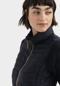 Marineblå quiltet vest med høj krave, der har en frontlynlås og brune læderdetaljer. Letvægtsmateriale med en struktureret overflade.