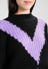Schwarzer Strickpullover mit einem markanten lila V-Muster auf der Vorderseite. Der Stoff wirkt strukturiert mit grobem Strick.