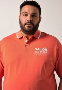 Korálová polo košile s bílými akcenty na límci. Na levé části hrudníku je vyšitý text "SAILOR 53.281534 M 2007". Měkká textilie.