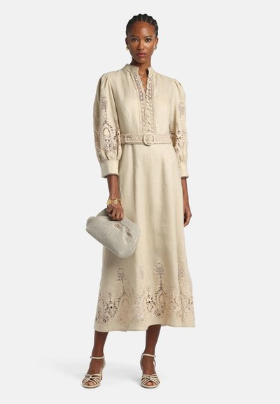 Vrouw die een beige midi-jurk draagt met pofmouwen, kanten details, een bijpassende riem, een gestructureerde clutch vasthoudt en open tenen hakken draagt.