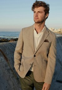 Blazer ajustado color marrón claro con un tejido texturizado, con un bolsillo en el pecho con un patrón, llevado sobre una camiseta polo crema y pantalones verdes.