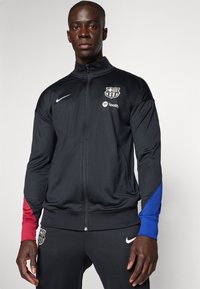 Sort Nike trackjakke med røde og blå detaljer, har en lynlås foran, Barcelona-logo og Spotify-branding på brystet.
