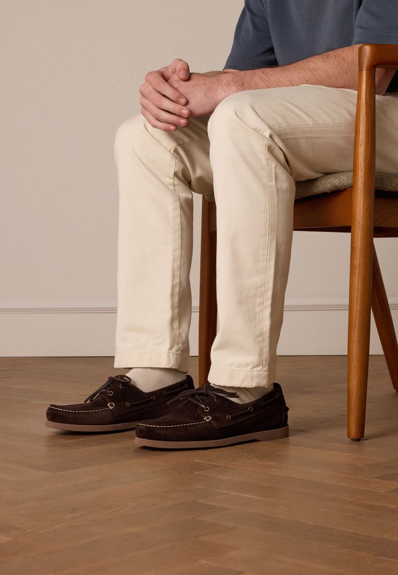 Bruine suède bootsschoenen met veters, gouden oogjes en witte stiksels, gedragen met lichte broeken, zittend op een houten vloer.
