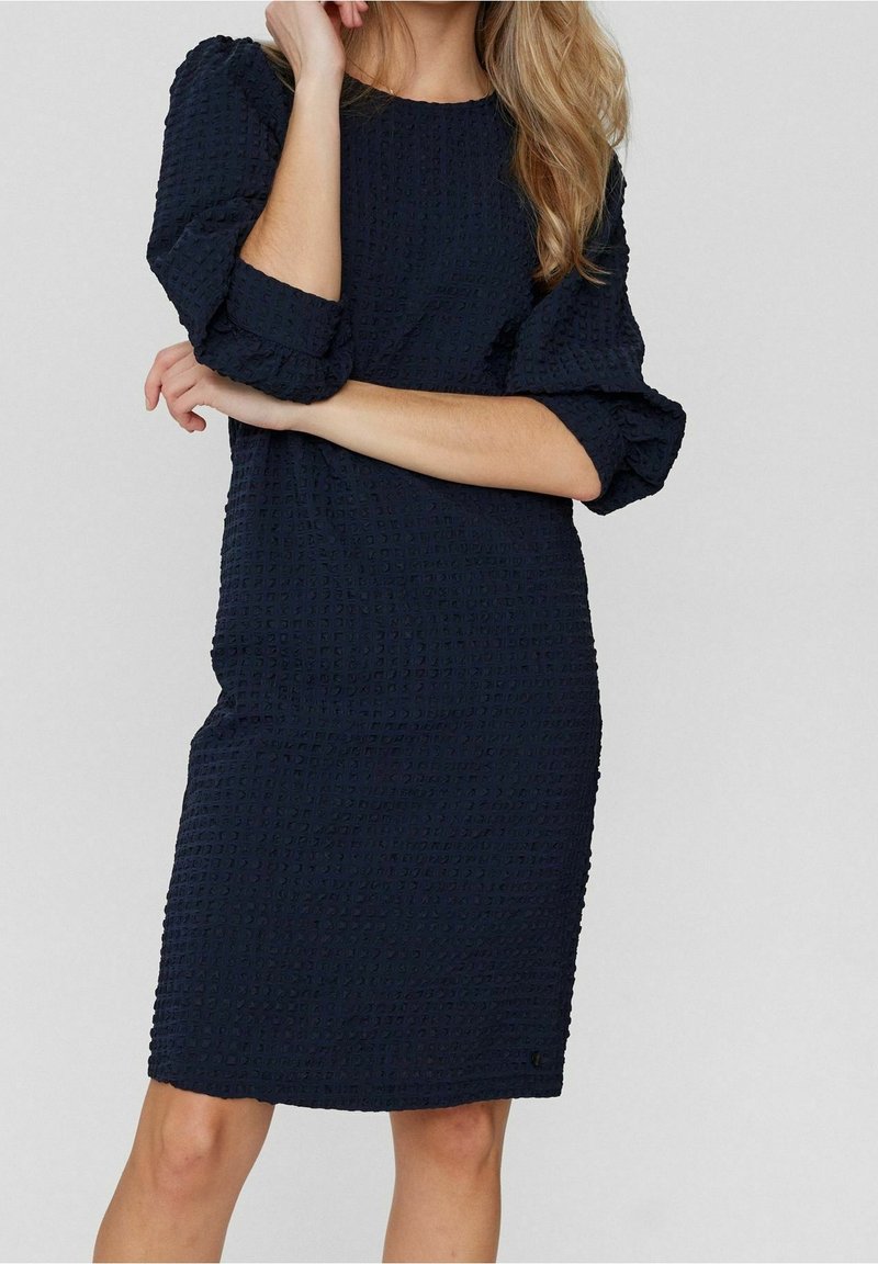 Femme portant une robe texturée bleu marine longueur genou avec des manches bouffantes trois-quarts, debout, les bras croisés et une main près du visage.
