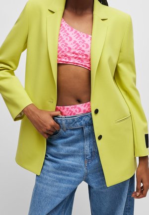 Person trägt limettengrünen Blazer über rosa gemustertem Croptop und hoch taillierten blauen Jeans, Hand in den Bund gesteckt.