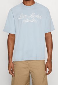 Světle modré bavlněné tričko s kulatým výstřihem a krátkými rukávy. Na přední straně má bílé vyšívané nápis "Low Lights Studios".