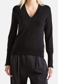 Kvinna som bär en svart långärmad V-halsad tröja inskjuten i löst sittande svarta byxor, stående mot en ljus bakgrund.