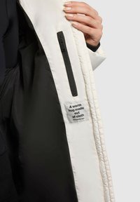 Fehér szigetelt kabát belseje, melyen látható egy fekete cipzáras zseb és egy címke szöveggel: "Egy meleg ölelés, ami szövetből készült." Érzéki anyag látható.