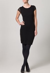 Černé přiléhavé šaty s krátkými rukávy a průhlednými akcenty na ramenech. Nosené s tmavými punčochami a lesklými černými lodičkami. Jednoduchý, elegantní design.