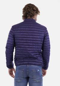 Giacca trapuntata viola con baffle stretti, colletto alto e una texture in nylon liscia. Indossata sopra dei jeans blu, con un'etichetta sulla tasca posteriore.