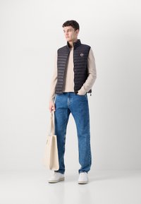 Gilet trapuntato nero su un maglione beige, abbinato a jeans blu e sneakers bianche. Una borsa tote crema è tenuta nella mano destra.