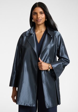 Donna con indosso un cappotto oversize lucido blu scuro, orecchini a cerchio dorati e un braccialetto d'oro, in piedi contro uno sfondo chiaro e semplice.