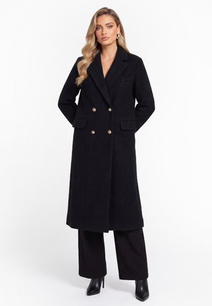 Woman wearing a long black double-breasted coat with gold buttons, black wide-leg pants, and black heeled ankle boots, standing with hands behind back.
