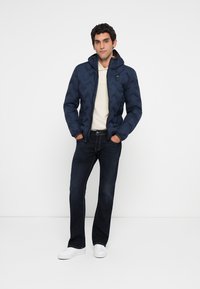 Bărbat aflat în picioare, cu mâinile în buzunare, purtând o jachetă matlasată albastru închis, un tricou polo alb, blugi negri și adidași albi, pe un fundal simplu.