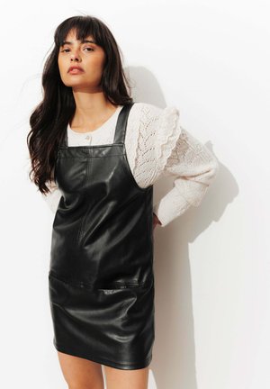 Robe en cuir noir avec un corsage ajusté et des bretelles réglables, associée à un pull en tricot blanc avec des accents en dentelle.