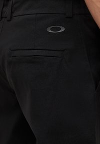 Κοντινό πλάνο μαύρου παντελονιού με πίσω τσέπη welt και ματ μαύρο οβάλ λογότυπο κοντά στην τσέπη στη δεξιά πλευρά.