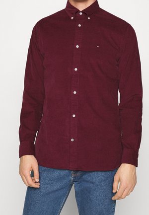 Mężczyzna ubrany w długaśną, zapinaną na guziki ciemnoczerwoną koszulę sztruksową i niebieskie dżinsy, stoi z luźno opuszczonymi ramionami.