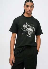 Svart bomulls T-skjorte med grafisk design som viser et øye, en hånd og en diamantform, aksentuert med hvite stjerner og kjededetaljer.