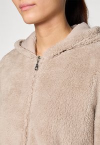 Veste en polaire beige avec capuche, dotée d'une fermeture éclair à l'avant, d'une texture douce et de détails de couture visibles. Design confortable et décontracté.