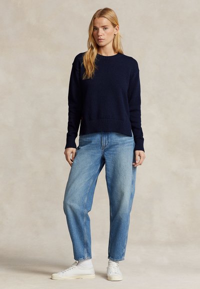 Tumši zils adīts džemperis ar apaļu kakla izgriezumu un ribota auduma apakšu, kopā ar gaismzilām vaļīgām džinsu biksēm un baltām sporta kurpēm.