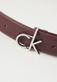 Cintura in pelle bordeaux con fibbia in metallo argentato a logo CK, superficie testurizzata e più fori di chiusura per la regolazione.